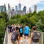 People walking on an elevated pathway surrounded by greenery, with a city skyline featuring skyscrapers in the background.