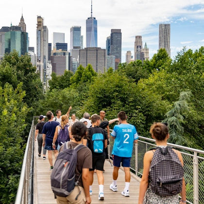 People walking on an elevated pathway surrounded by greenery, with a city skyline featuring skyscrapers in the background.