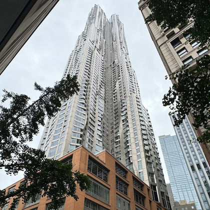 A tall, modern skyscraper with a unique, wavy design against a cloudy sky, surrounded by trees and other buildings.