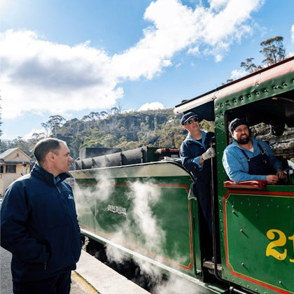 Two train operators stand beside and inside a green steam locomotive at a station, with a person observing nearby.