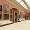 A grand room with ornate pink walls, framed paintings, white benches, and a glass ceiling, creating an opulent, gallery-like setting.