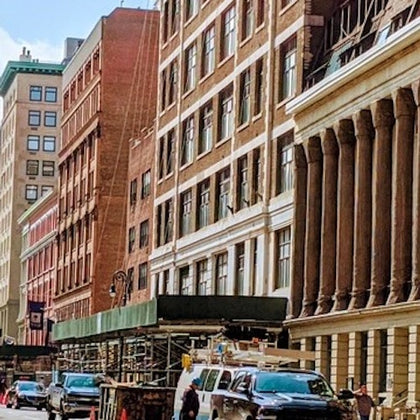 City street with brick buildings, a row of columns, parked vehicles, and scaffolding along the sidewalks.