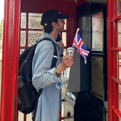 A person in a denim jacket and backpack holds a small Union Jack flag inside a classic red phone booth.