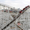 Red roller coaster car descending a wooden track with a city skyline in the background under a cloudy sky.