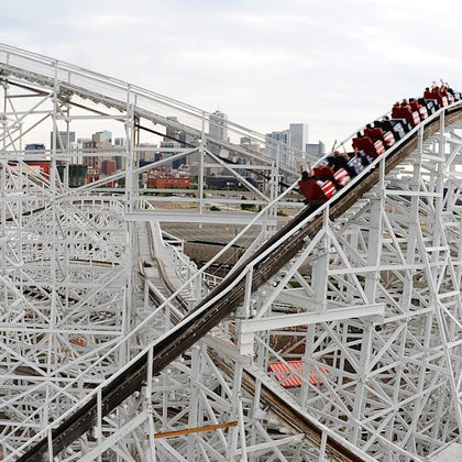 Red roller coaster car descending a wooden track with a city skyline in the background under a cloudy sky.