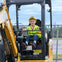 A young child wearing a yellow vest and hard hat sits in the operator's seat of a small excavator.
