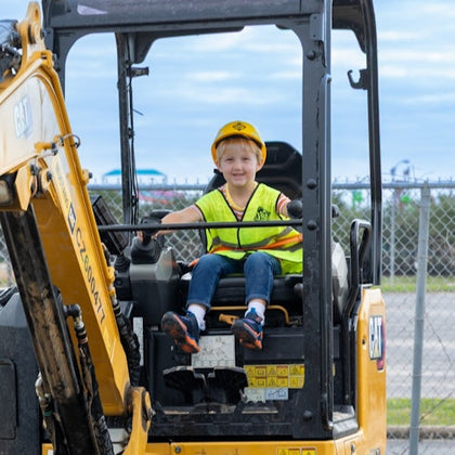 A young child wearing a yellow vest and hard hat sits in the operator's seat of a small excavator.