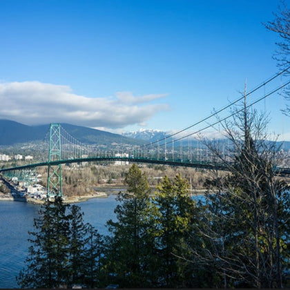 A suspension bridge spans over a body of water, surrounded by trees and mountains under a clear blue sky.