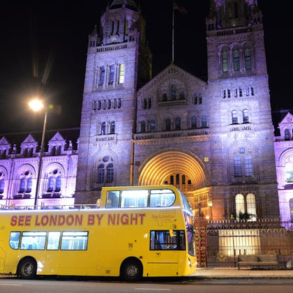 A yellow double-decker bus with "See London by Night" written on it, parked in front of an illuminated historic building at night.