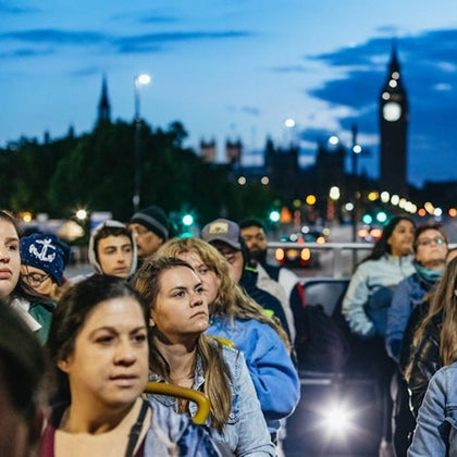 A group of people outdoors during twilight, with the silhouette of Big Ben and Westminster Palace in the background.