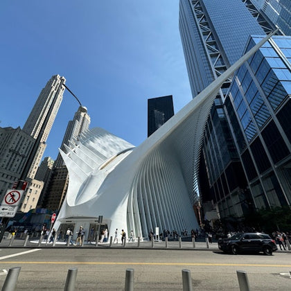 A modern white architectural structure with curved features stands amidst tall buildings, with people and cars nearby.