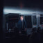 A man stands on a stage with a microphone in a dimly lit room in front of an audience.