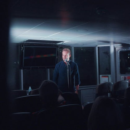 A man stands on a stage with a microphone in a dimly lit room in front of an audience.