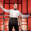 Man in a white shirt and bow tie with arms outstretched, singing passionately on stage with a red and black background.