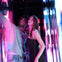 Two people smiling inside a brightly lit, colorful space with reflective surfaces and vivid light patterns.