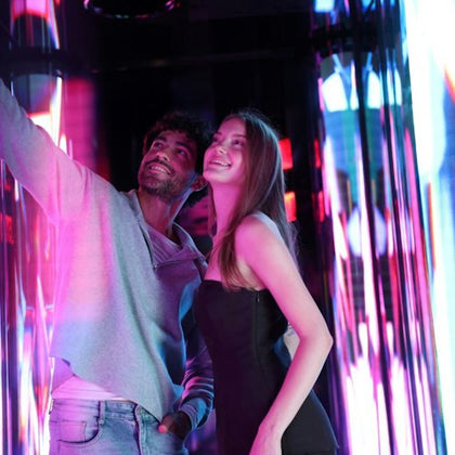 Two people smiling inside a brightly lit, colorful space with reflective surfaces and vivid light patterns.