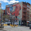Urban street scene with a large mural of a red-clad figure on a building, surrounded by graffiti and outdoor seating.