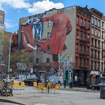 Urban street scene with a large mural of a red-clad figure on a building, surrounded by graffiti and outdoor seating.