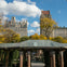 Buildings and trees in the background with a pavilion featuring brick columns and a green roof in the foreground. Blue sky with clouds.