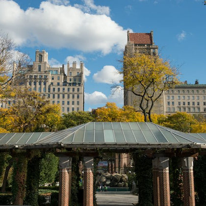 Buildings and trees in the background with a pavilion featuring brick columns and a green roof in the foreground. Blue sky with clouds.