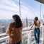 People standing on a glass observation deck overlooking a cityscape with prominent buildings under a clear sky.