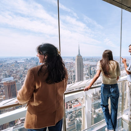 People standing on a glass observation deck overlooking a cityscape with prominent buildings under a clear sky.