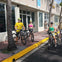 Three people wearing helmets on rental bikes on a sunny sidewalk outside a building with large windows and palm trees.