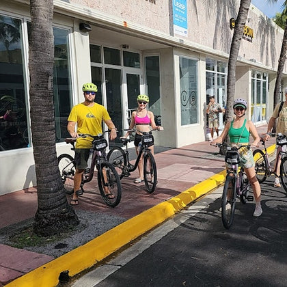 Three people wearing helmets on rental bikes on a sunny sidewalk outside a building with large windows and palm trees.