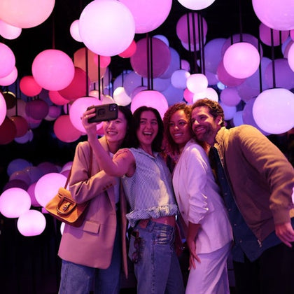 Four people taking a selfie surrounded by colorful glowing spheres in a vibrant, atmospheric space.
