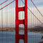 Close-up view of a red suspension bridge tower with cable lines, with a cityscape and water in the background.