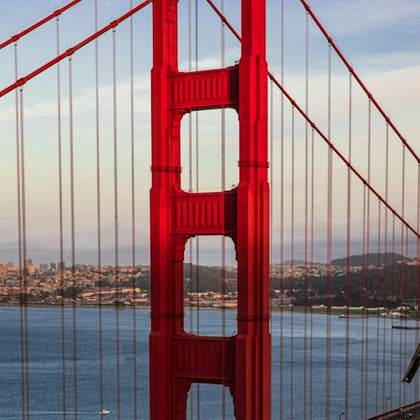 Close-up view of a red suspension bridge tower with cable lines, with a cityscape and water in the background.
