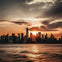 A city skyline at sunset with the sun setting behind tall buildings, reflecting light on a calm body of water in the foreground.