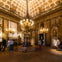 An ornate room with chandeliers, golden decorations, and statues. Visitors are observing the exhibits on display.
