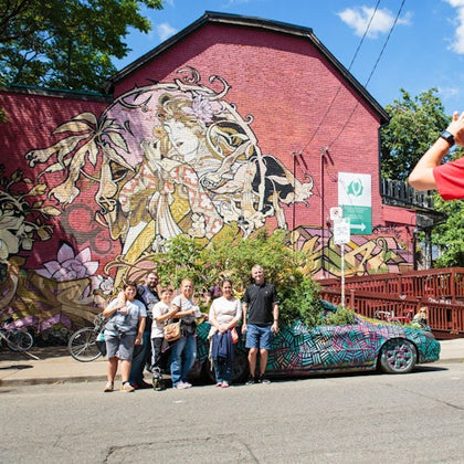 A group of people pose near a colorful car in front of a building with vibrant graffiti art. One person stands off to the side.