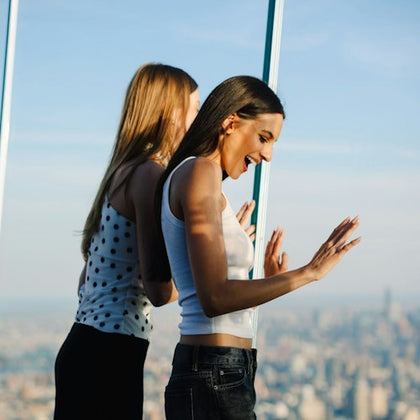 Two women standing near a glass wall, one smiling and gesturing excitedly, overlooking a cityscape in the background.