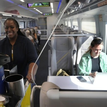 A smiling staff member serves refreshments on a modern train; passengers are seated, one working on a laptop.