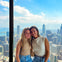 Two smiling individuals sit in front of a large window with a cityscape and a body of water visible in the background.