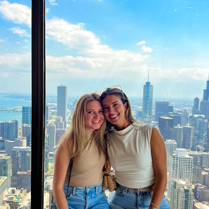 Two smiling individuals sit in front of a large window with a cityscape and a body of water visible in the background.