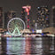 Nighttime cityscape featuring a ferris wheel, illuminated skyscrapers, and reflections on water.