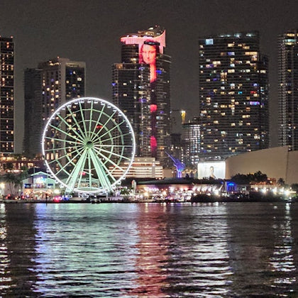Nighttime cityscape featuring a ferris wheel, illuminated skyscrapers, and reflections on water.