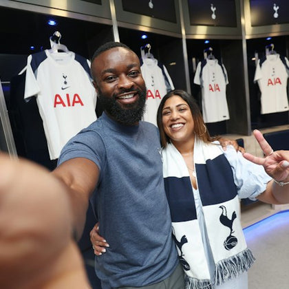 A smiling man and woman take a selfie in a locker room with Tottenham Hotspur jerseys hanging in the background.