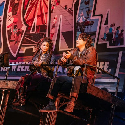 Two people sitting on a raised platform in front of a colorful backdrop with carnival illustrations and large text.