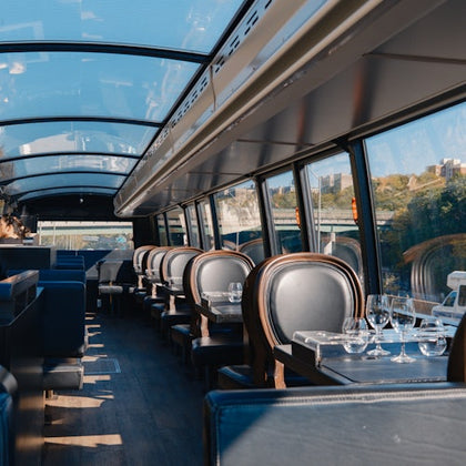 Interior of an empty, glass-roofed train dining car with set tables, chairs, and scenic views outside the window.