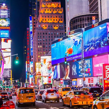 Bustling Times Square at night with bright advertising billboards, heavy traffic, yellow taxis, and crowded sidewalks.