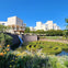 Stone buildings with terraces overlook a manicured garden with a water feature and maze-like hedges under a clear blue sky.
