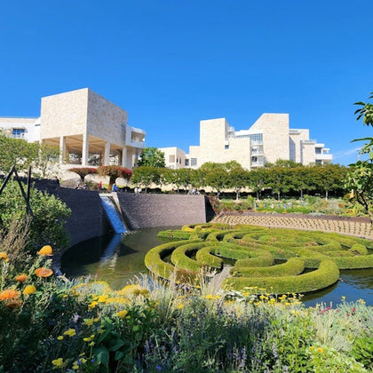 Stone buildings with terraces overlook a manicured garden with a water feature and maze-like hedges under a clear blue sky.