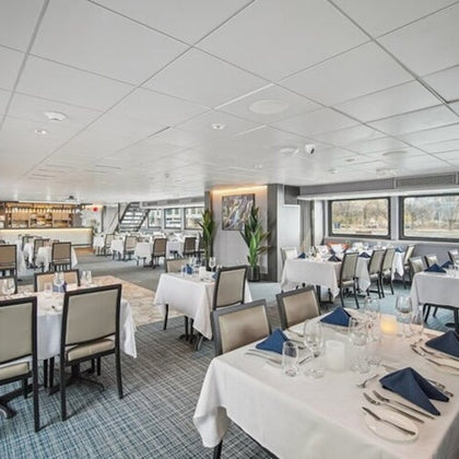 A modern restaurant interior with neatly arranged tables, white tablecloths, and blue napkins, featuring large windows and a bar area.