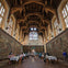 A grand hall with ornate wooden ceiling, stained glass windows, large tapestries on the walls, and people seated at tables.