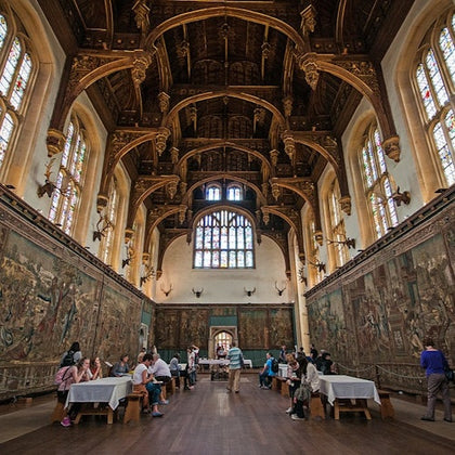 A grand hall with ornate wooden ceiling, stained glass windows, large tapestries on the walls, and people seated at tables.