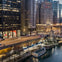 Elevated view of a cityscape at dusk showing tall buildings, a busy road, illuminated streetlights, and boats docked on a river.